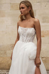 A woman wearing a strapless white wedding dress with a lace corset-style bodice and a voluminous tulle skirt, standing against a light stone wall.
