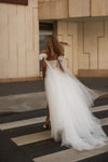 A woman wearing a voluminous white tulle wedding dress with puff sleeves and a backless open-back design, walking across a crosswalk outdoors in front of a beige stone building.