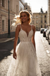 A woman wearing a white lace wedding gown stands on a sunlit city street, with buildings and parked cars blurred in the background.