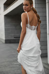 A woman wearing a white spaghetti-strap maxi dress with a lace-trimmed open back, photographed outdoors against a gray building wall.