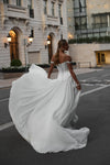 A woman wearing a white off-the-shoulder wedding gown with a structured corset-style bodice and a voluminous flowing skirt, photographed outdoors on a city street with buildings in the background.