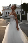 A woman wearing a strapless white wedding gown with a fitted corset-style bodice and full tulle skirt, standing outdoors on a street with a building in the background.