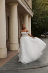A woman wearing a voluminous off-the-shoulder white wedding gown stands on a stone walkway in front of a building with large cream columns, holding out one side of the skirt to show its full volume.