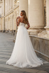 A woman wearing a white off-the-shoulder wedding gown with delicate chain straps and a full tulle skirt, standing on a stone walkway beside a classical building with large columns.