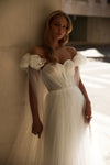 A woman wearing a voluminous off-the-shoulder white wedding gown with ruffled sleeves, leaning against a textured wall in a sunlit architectural setting.