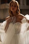 A woman wearing a strapless white tulle wedding dress with dramatic puffed sleeves and a sheer overlay, photographed in warm natural light against a softly blurred outdoor background.