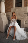 A woman wearing a strapless white wedding dress with long lace sleeves sits on stone steps in front of a brick building. The dress has a fitted bodice, sheer lace sleeves, and a layered tulle skirt with a train.