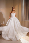 A woman models a strapless white ball gown wedding dress with a structured bodice and a voluminous flowing skirt in a warmly lit indoor setting.