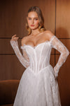 A woman modeling an off-the-shoulder white lace wedding gown with a fitted corset-style bodice and long sheer lace sleeves, posed against a warm brown wood-paneled background.