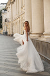 A woman wearing a flowing white wedding gown stands on a stone walkway beside a large classical building, with the dress billowing in the wind.