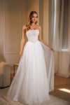 A woman modeling a strapless white wedding gown with a structured corset-style bodice and a full tulle skirt, standing indoors near sheer curtains and warm ambient light.