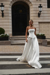A woman wearing a strapless white wedding gown stands on a crosswalk in front of a grand stone building with a dark arched doorway.