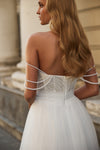 A woman shown from the back wearing a white strapless wedding dress with pearl-like draped straps and a heavily embellished beaded bodice, photographed outdoors against a soft beige stone background.