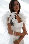 A woman wearing a white sleeveless dress with a large white feathered angel wing accessory attached to her shoulders, posed against a bright white background.