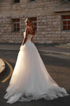 A woman wearing a strapless white wedding gown with a fitted lace bodice and a full tulle skirt, standing outdoors in front of a stone building.
