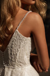 Close-up back view of a woman wearing a white beaded wedding dress with pearl-like embellished straps and a beaded waistband, against a dark background.