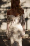 An off-the-shoulder lace wedding gown with long sheer lace sleeves and a fitted mermaid silhouette, shown on a model in warm natural light against a softly blurred outdoor background.