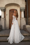 A woman wearing a white off-the-shoulder lace wedding gown with long sheer sleeves and a full skirt, standing on stone steps in front of a building with a wooden door and columns.