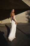 A woman wearing a flowing white sleeveless dress stands outdoors under a concrete structure, captured in warm sunlight with strong shadows.