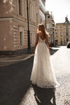 A woman wearing a white lace wedding gown stands on a sunlit city street, photographed from behind as she looks toward the buildings.