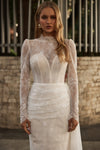 A woman wearing a white lace wedding dress with long sheer sleeves, a high mock neck, and a fitted corset-style bodice, standing outdoors against a stone wall and metal fence.