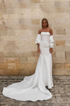 A woman wearing an off-the-shoulder white wedding gown with dramatic puffed sleeves and a long train, standing against a beige stone wall and cobblestone ground.
