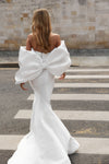 A woman viewed from behind wearing a white off-the-shoulder mermaid gown with oversized dramatic bow details on the back, standing on a crosswalk in front of a stone wall.