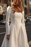 A woman wearing a white long-sleeve dress with a square neckline and structured skirt, photographed outdoors on a city street with blurred buildings and cars in the background.