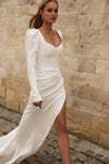 A woman wearing a fitted white long-sleeve wedding dress with a deep V-neckline, puffed shoulders, and a high side slit, standing outdoors against a beige stone wall.