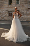 A woman modeling a strapless white wedding gown with a structured corset-style bodice and a full tulle skirt, standing outdoors on a street in front of a stone building.
