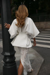 A woman viewed from behind wearing a white short-sleeve peplum-style top and white lace pants, standing on a city sidewalk next to a black lamppost.
