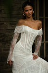 A woman wearing an off-the-shoulder white lace wedding gown with long sheer sleeves and a fitted corset-style bodice, posed against a dark brick wall and window.