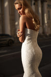 A woman wearing a strapless fitted white lace dress stands outdoors on a sunlit city street, photographed from the back and side.