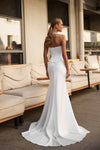 A woman wearing a strapless white wedding gown with a fitted corset-style bodice and a long train, photographed from the back in an outdoor patio setting with beige seating.