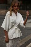 A woman wearing a white wrap-style cape jacket with a tie waist, paired with a white lace skirt, standing on a city street.