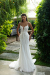A woman modeling a white sleeveless wedding gown with thin straps, a deep V neckline, and a fitted mermaid silhouette, standing outdoors on a stone path surrounded by green foliage.