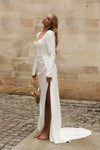 A woman wearing a long-sleeve white wedding dress with a thigh-high slit, standing barefoot outdoors against a beige stone wall and cobblestone ground.