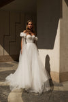 A woman wearing a voluminous off-the-shoulder white wedding gown with a fitted bodice and flowing tulle skirt, standing in a sunlit architectural setting with strong shadows.