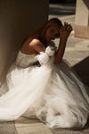 A woman in a voluminous white tulle wedding gown sits on stone steps in dramatic, high-contrast sunlight and shadow.