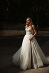 A woman wearing a strapless white wedding gown with a fitted lace bodice and a full tulle skirt, standing outdoors on a paved street with dark trees and shadows in the background.