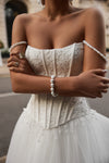 A close-up of a woman wearing a strapless white lace wedding gown with a fitted corset-style bodice, tulle skirt, and pearl-beaded fringe details. She is also wearing pearl bracelets and rings, with a softly blurred outdoor architectural background.