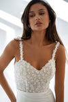 A woman modeling a white lace bridal top with floral embroidery and beaded details, paired with a white skirt, set against a bright, modern white background.