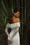 A woman wearing an off-the-shoulder white lace dress with long sheer lace sleeves, standing outdoors beside a brick wall and tree trunk.