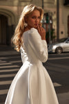 A woman wearing a white long-sleeve wedding dress with structured shoulders and a full skirt, photographed outdoors on a city street with blurred buildings and a car in the background.