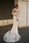 A woman wearing a long-sleeve lace wedding gown with an open back and dramatic train, standing outdoors against a beige wall and dark shuttered window.