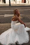 A woman wearing a strapless white lace wedding gown sits on a city street curb, with the dress spread out around her.