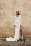 A woman wearing a long-sleeved white wedding gown with a high thigh slit and train, standing against a beige stone wall on a cobblestone surface.