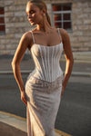 A woman wearing a white lace corset-style dress with thin straps and a fitted silhouette, standing outdoors in front of a stone building at warm golden-hour light.