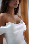 Close-up of a woman wearing a white off-the-shoulder wedding dress with a beaded sweetheart neckline and a wide draped sleeve.