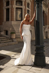A woman modeling a strapless white wedding gown with a structured bodice and fitted mermaid-style skirt, standing outdoors beside a dark lamppost in front of a stone building.